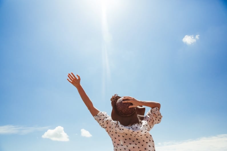 A woman with a shirt and hat holds her arms up towards the sun shining in a blue sky
