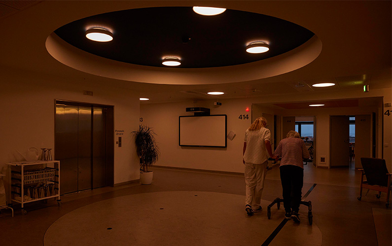 Nurse and patient with a walker stroll down a corridor arm in arm in a hospital unit with circadian lighting