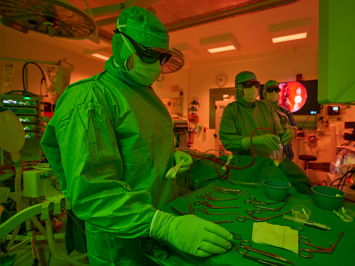 Operations­sjuksköterskan tittar på instrument medan kirurgen står i bakgrunden och utför operation i ergonomisk ljus