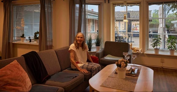 Ældre kvinde sidder smilende på en sofa i en stue og ser sig til siden
