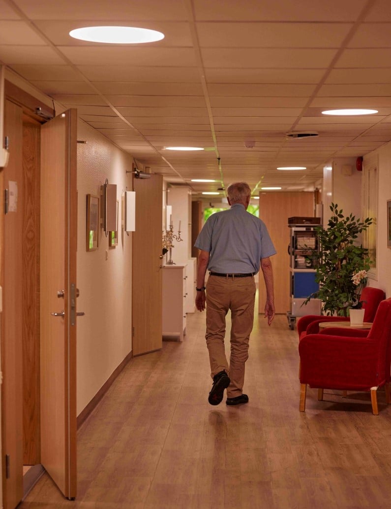 An older man walks down a long hall with circadian lighting