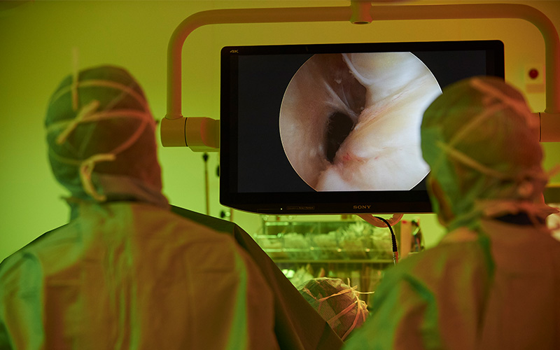 To kirurger i uniform står med ryggen til i ergonomisk lys og kigger på en monitor imens de udfører kikkertkirurgi