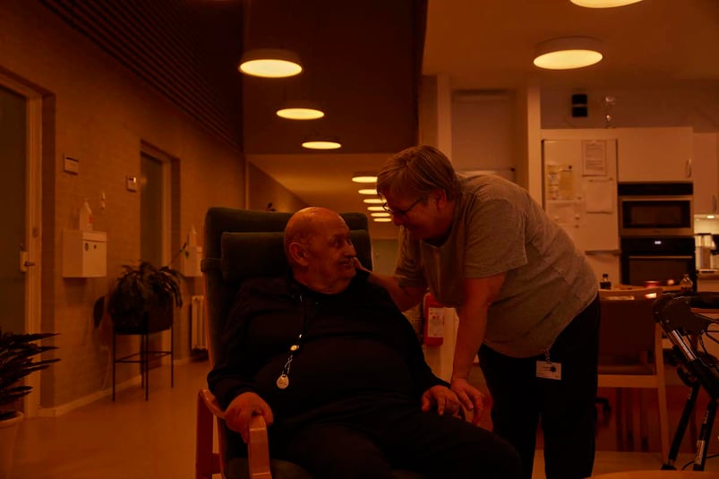 Medarbejder taler med en smilende ældre borger i døgnrytmelyset på fællesstuen på plejehjemmet Bauneparken